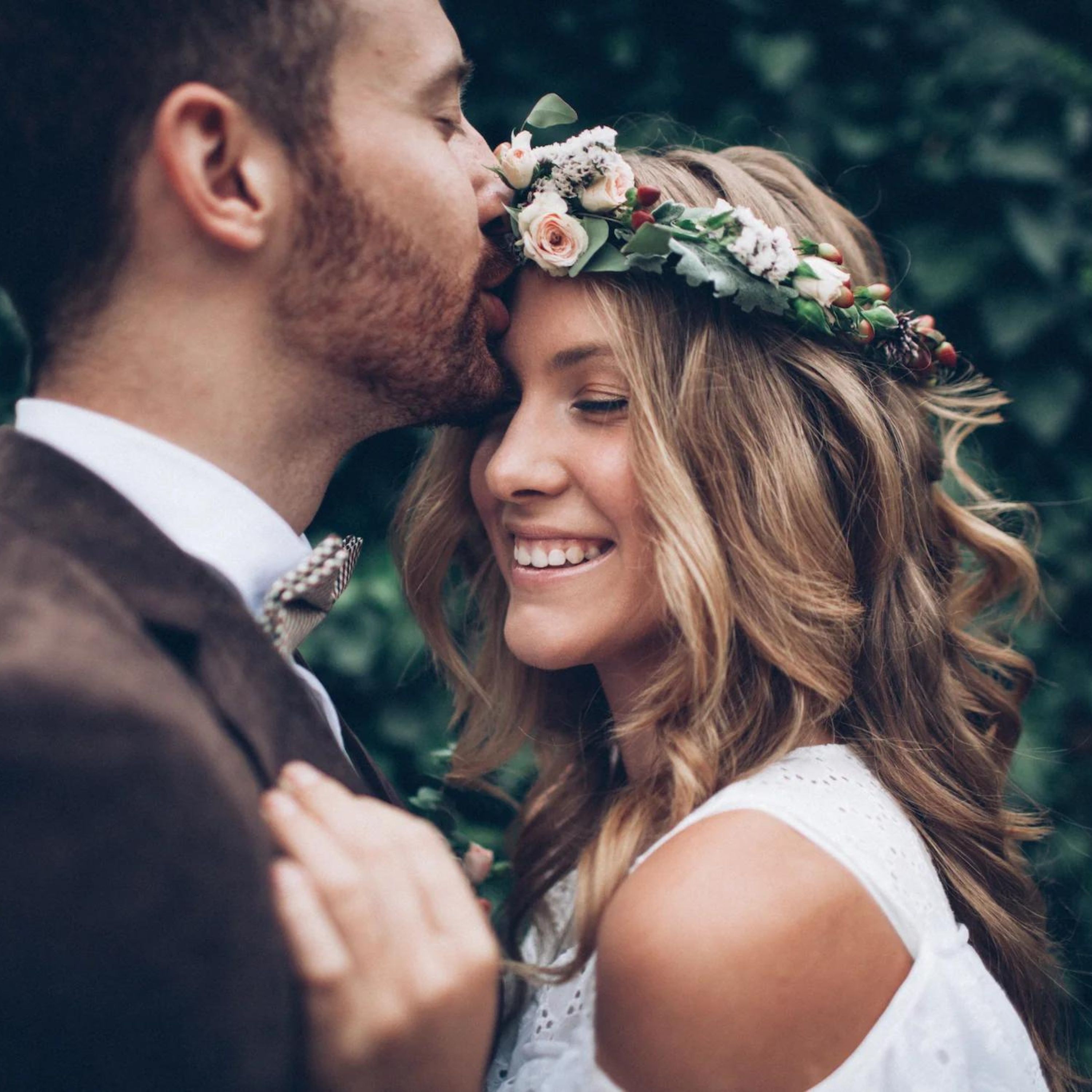 Ein Mann und eine Frau stehen nah beieinander, der Mann gibt der Frau einen Kuss auf die Stirn. Sie feiern ihre Hochzeit.