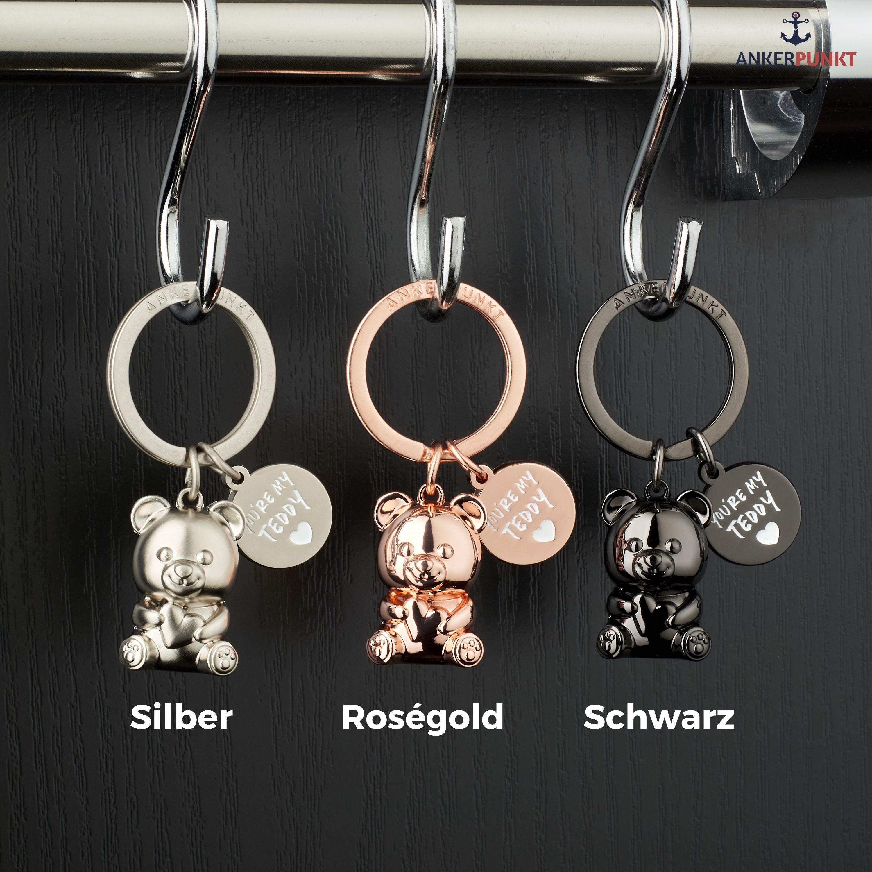 Drei Teddybären-Anhänger hängen an drei Schlüsselhaken vor einer schwarzen Wand.