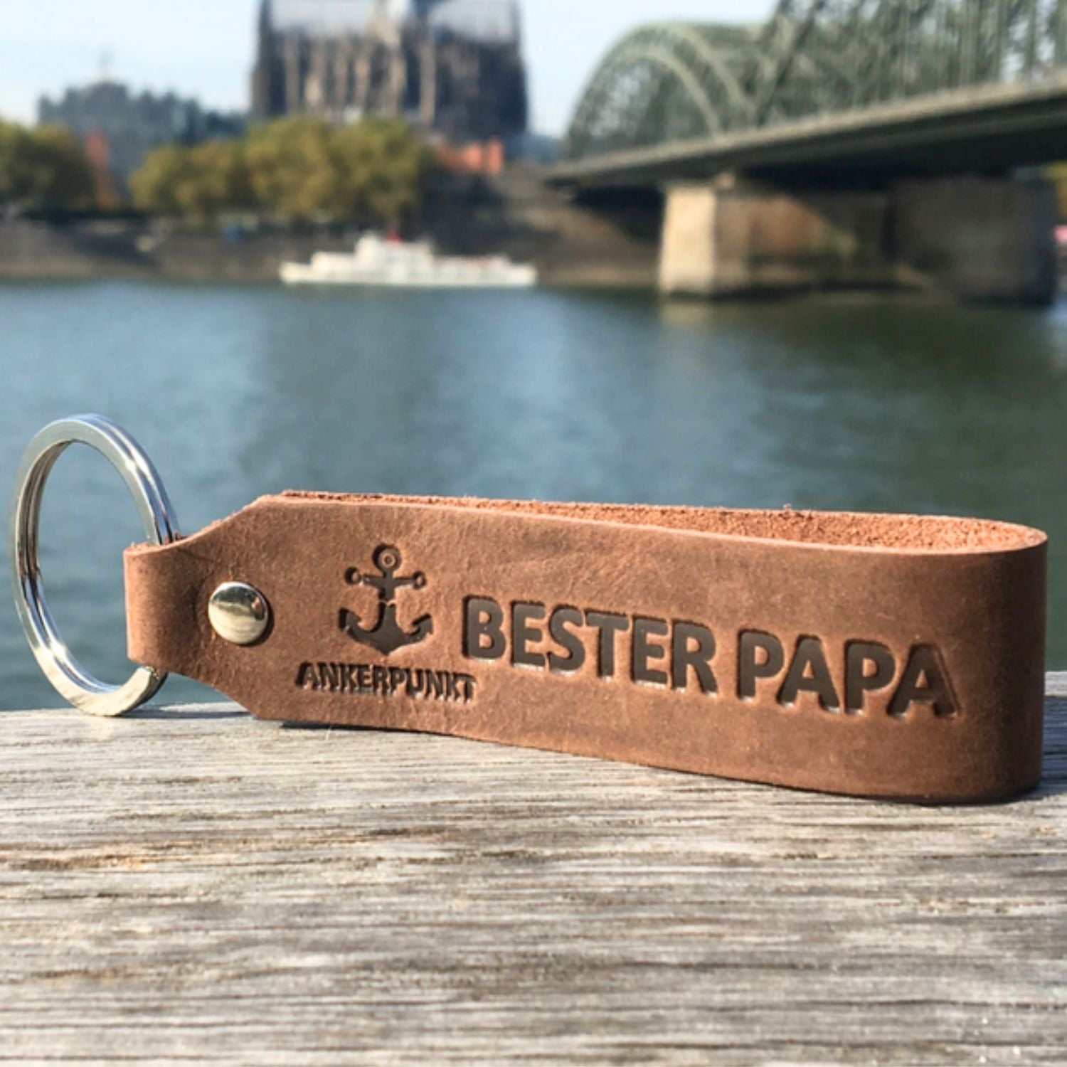 Dark brown leather keychain with "Bester Papa" engraving, river and bridge background.