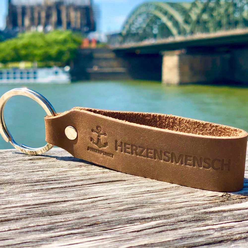 Herzensmensch Schlüsselanhänger aus dunkelbraunem Leder auf Holzoberfläche vor Flusskulisse.