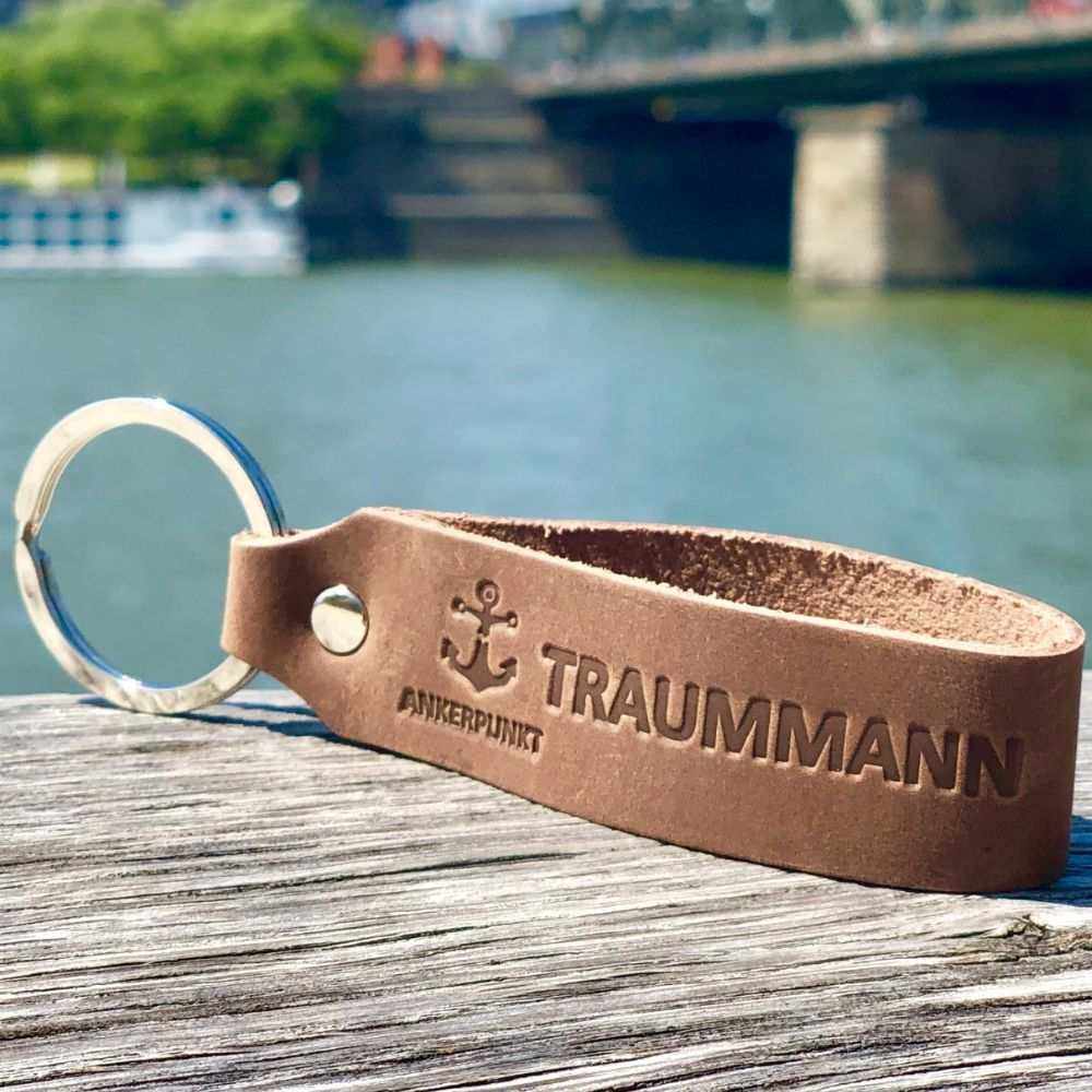 Traummann Schlüsselanhänger aus dunkelbraunem Leder auf Holztisch vor Flusskulisse.