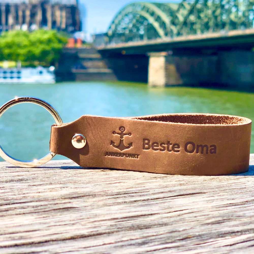 Leather keychain with "Beste Oma" engraving, made in Germany, lying on a wooden surface by a river.