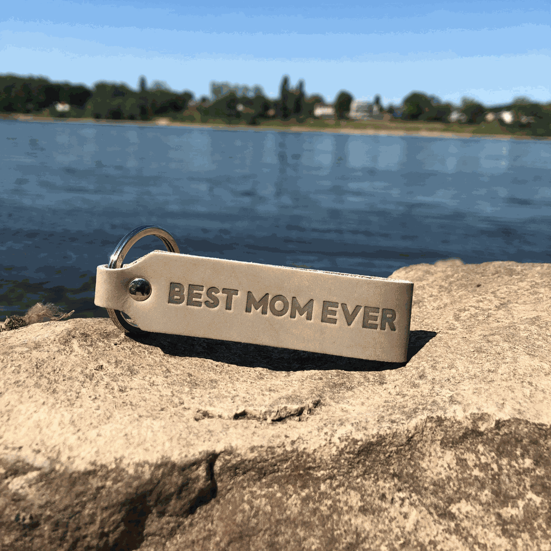Leder-Schlüsselanhänger "Best Mom Ever" in Grau auf Felsen am See.