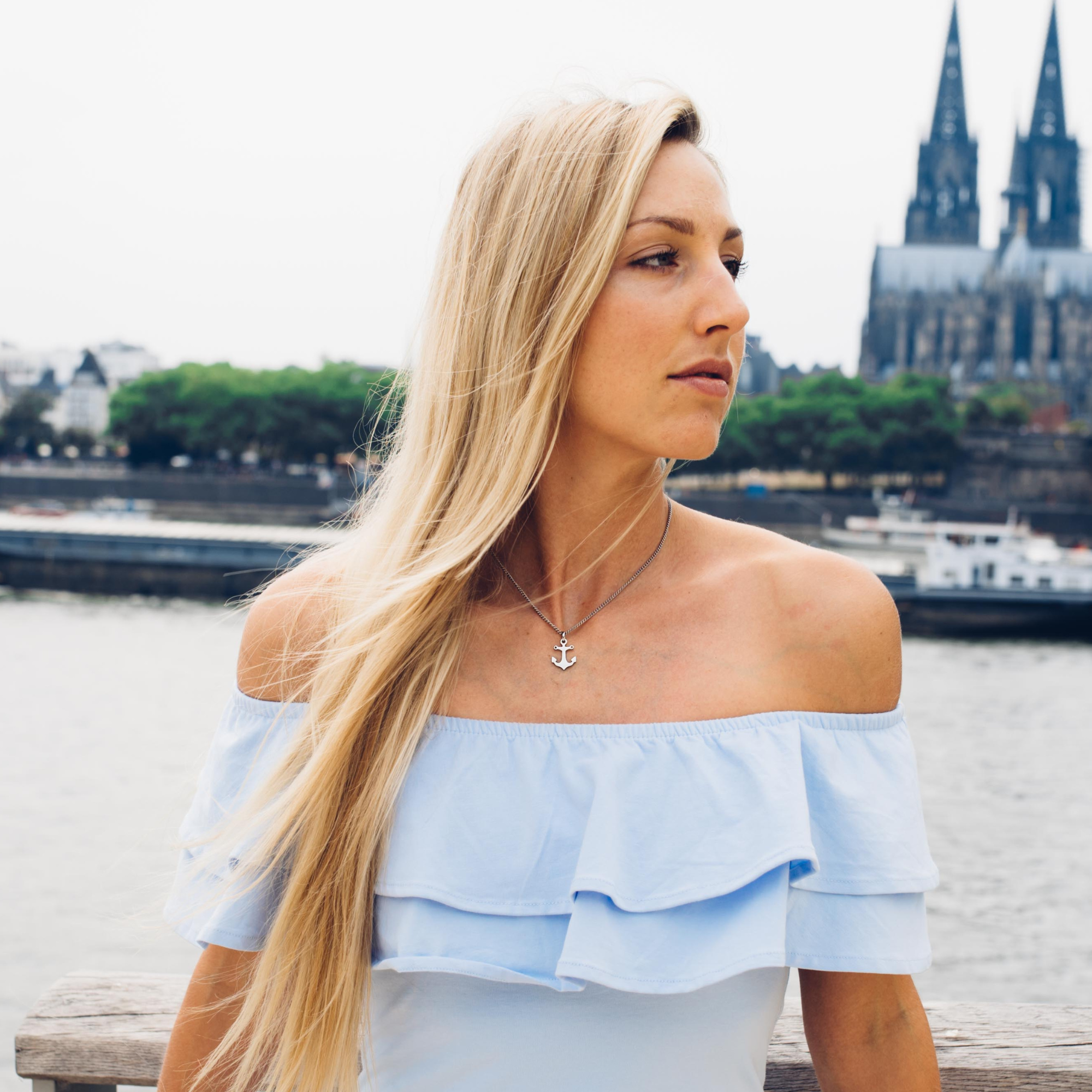 Eine blonde Frau steht am Rhein und hat eine Ankerpunkt-Kette um den Hals. Im Hintergrund ist der Kölner Dom zu sehen.